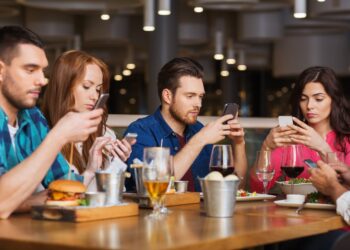 Junge Menschen sitzen zusammen beim Essen und schauen auf ihr Smartphone.
