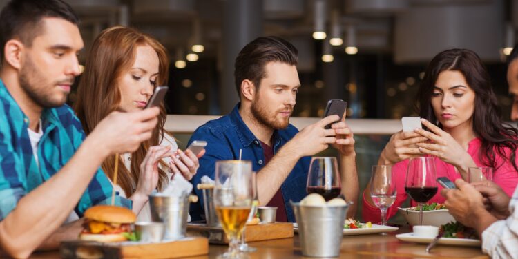 Junge Menschen sitzen zusammen beim Essen und schauen auf ihr Smartphone.