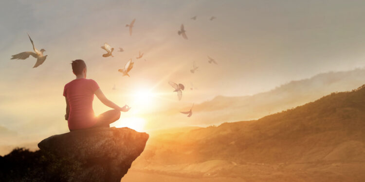 Eine Frau sitzt auf einem Felsvorsprung und meditiert, während Vögel über ihr fliegen und die Sonne am Himmel steht.