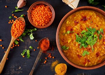 Ein Teller mit Linsen-Curry ist neben einigen Zutaten und Gewürzen auf einer schwarzen Oberfläche drapiert.