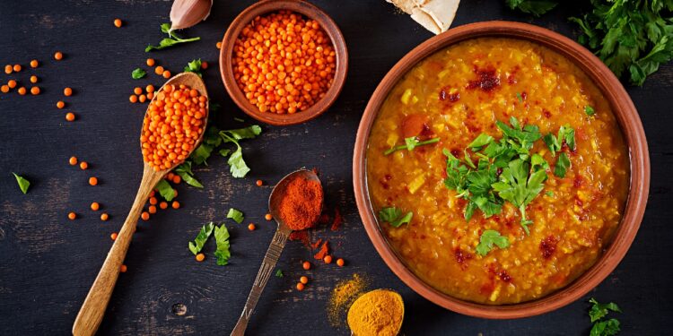 Ein Teller mit Linsen-Curry ist neben einigen Zutaten und Gewürzen auf einer schwarzen Oberfläche drapiert.