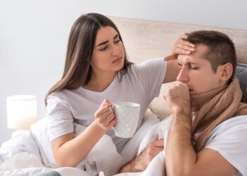 Ein Mann mit einem Schal um den Hals liegt im Bett. Eine Frau liegt daneben und fühlt mit einer Hand seine Stirn und reicht ihm mit der anderen eine Tasse.
