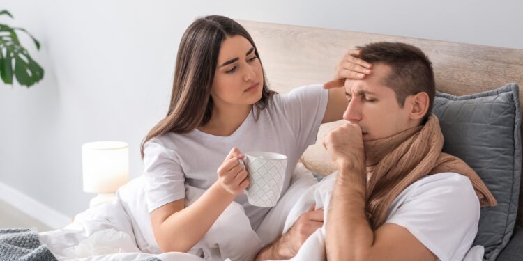 Ein Mann mit einem Schal um den Hals liegt im Bett. Eine Frau liegt daneben und fühlt mit einer Hand seine Stirn und reicht ihm mit der anderen eine Tasse.