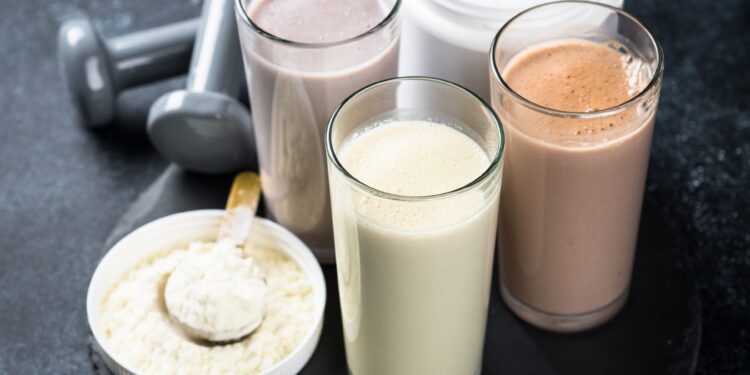 Verschiedene Proteinshakes in Gläsern, neben einem Eiweißpulver und Hanteln