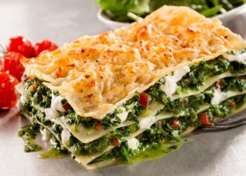 Ein Stück Spinat-Lasagne ist neben einigen Tomaten auf einer weißen Oberfläche angerichtet.