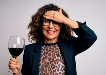 Eine Frau hält in einer Hand ein Glas Rotwein in der Hand und fasst dich mit der anderen Hand auf die Stirn.