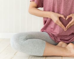 Eine Person sitzt im Schneidersitz auf den Boden und formt mit beiden Händen ein Herz vor dem Bauch.