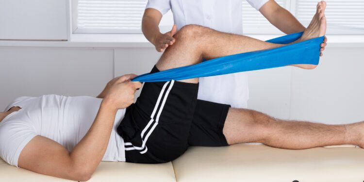 Physiotherapeut steht neben einer Behandlungsliege und unterstützt den darauf liegenden Patienten bei einer Übung mit dem Flexiband