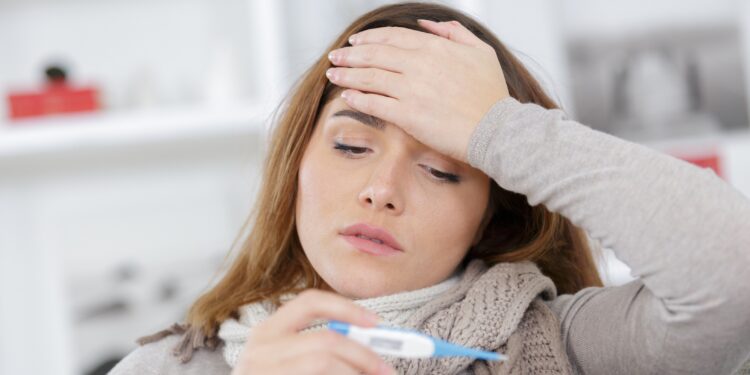 Frau mit Halstuch hält mit einer Hand ihre Stirn während sie in der anderen ein Fieberthermometer hält