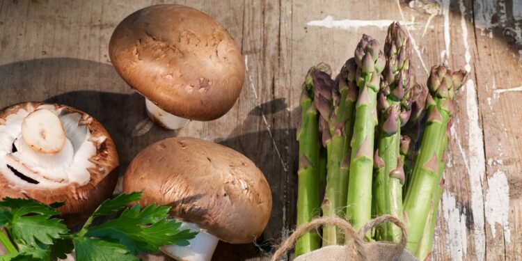 Ein Bund Spargel und einige Champignons liegen auf einer hölzernen Oberfläche.