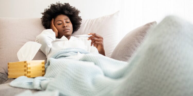 Frau liegt mit Decke und Taschentüchern auf dem Sofa, sie hält sich den Kopf und schaut auf ein Fieberthermometer