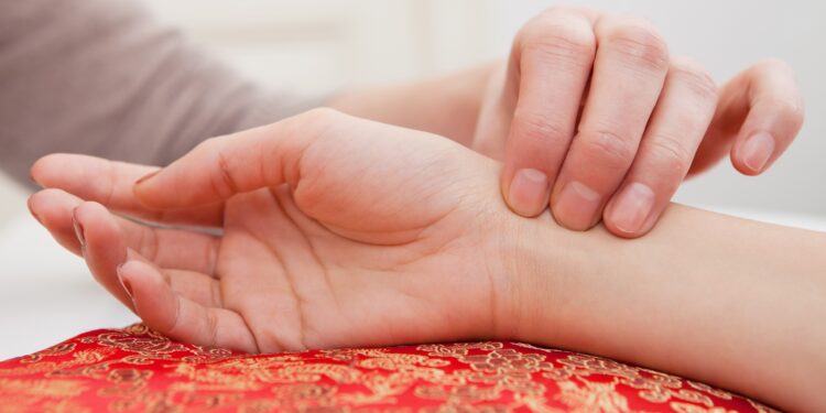 Behandler fühlt mit den Fingern den Puls am Handgelenk einer anderen Person, deren Hand auf einem rotgemusterten Kissen liegt