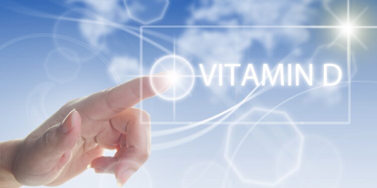 Finger zeigt auf den Schriftzug "Vitamin D" vor blauem Himmel mit strahlender Sonne.