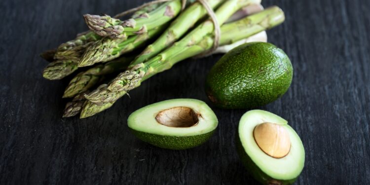 Ein Bund Spargel und ein paar Avocado liegen auf einer dunklen Oberfläche.