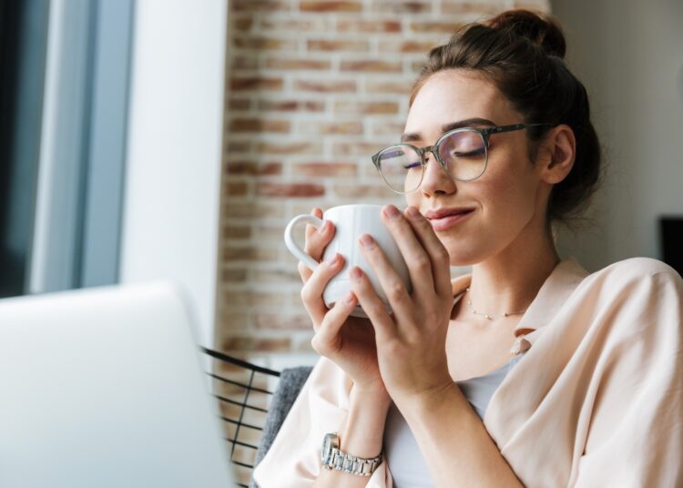 Frau sitzt auf dem Sofa und hält mit beiden Händen eine Tasse Kaffee.