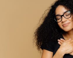 Mensch mit krausen dunklen Haaren und Brille lächelt und verschränkt die Hände über dem Herzen vor beigefarbenem Hintergrund