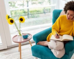 Mensch in gelbem Pullover sitzt in grünem Sessel und schreibt in ein Journal, auf einem Tisch daneben steht eine Vase mit Sonnenblumen