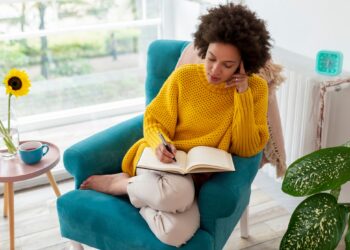 Mensch in gelbem Pullover sitzt in grünem Sessel und schreibt in ein Journal, auf einem Tisch daneben steht eine Vase mit Sonnenblumen