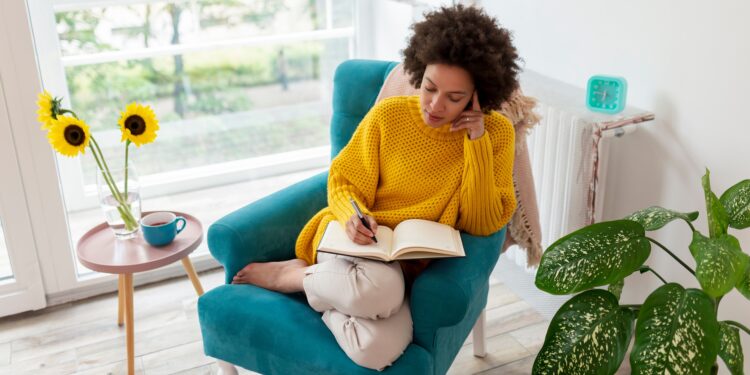 Mensch in gelbem Pullover sitzt in grünem Sessel und schreibt in ein Journal, auf einem Tisch daneben steht eine Vase mit Sonnenblumen