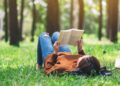Mensch in Jeans und Pulli liegt auf Wiese mit Bäumen und liest ein gedrucktes Buch