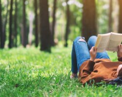 Mensch in Jeans und Pulli liegt auf Wiese mit Bäumen und liest ein gedrucktes Buch