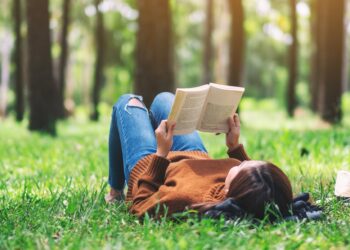 Mensch in Jeans und Pulli liegt auf Wiese mit Bäumen und liest ein gedrucktes Buch