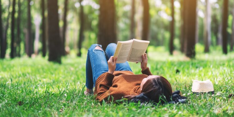 Mensch in Jeans und Pulli liegt auf Wiese mit Bäumen und liest ein gedrucktes Buch