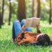 Mensch in Jeans und Pulli liegt auf Wiese mit Bäumen und liest ein gedrucktes Buch