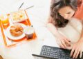 Frau liegt auf dem Bauch im Bett und tippt auf dem Laptop neben ihr steht ein Tablett mit frischem Orangensaft Croissant und Kaffee