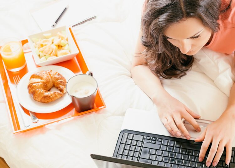 Frau liegt auf dem Bauch im Bett und tippt auf dem Laptop neben ihr steht ein Tablett mit frischem Orangensaft Croissant und Kaffee