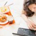 Frau liegt auf dem Bauch im Bett und tippt auf dem Laptop neben ihr steht ein Tablett mit frischem Orangensaft Croissant und Kaffee